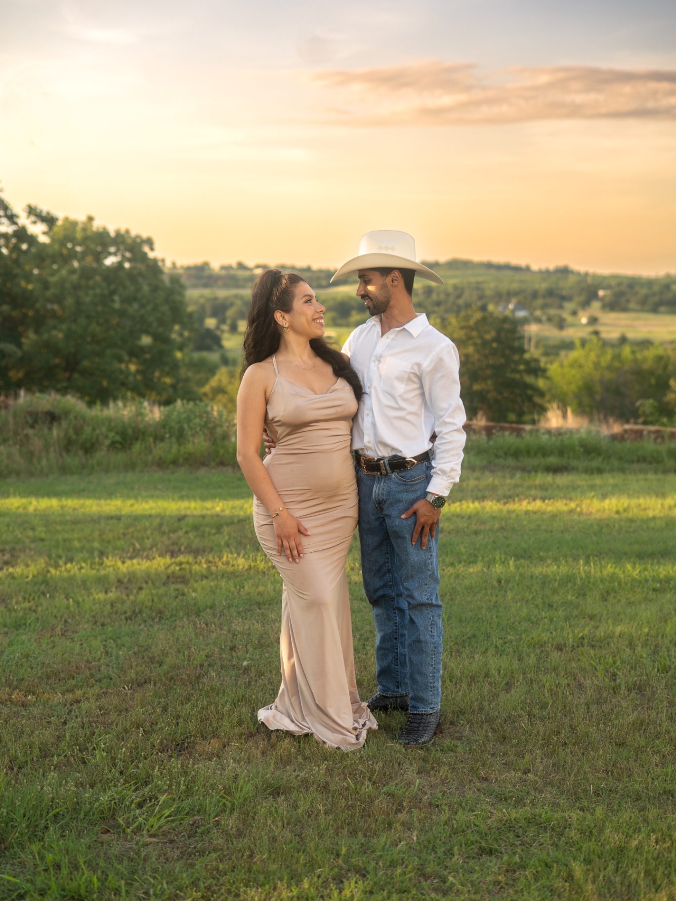 A couple at sunset in open green hills; she wears a champagne gown, he wears a white shirt and hat.