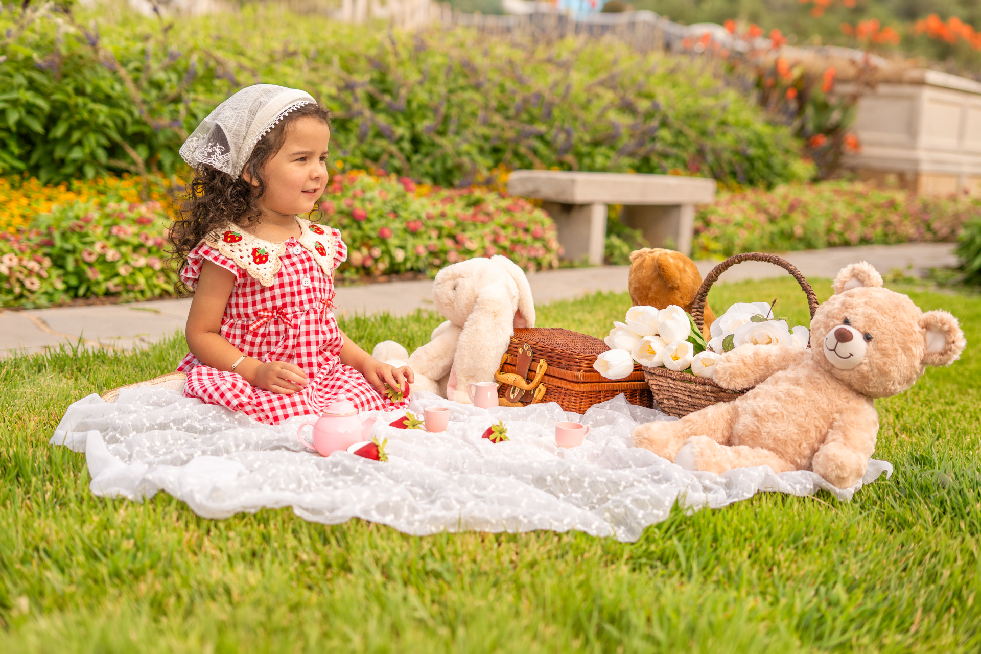 A young girl in a red gingham dress and lace headscarf at a garden picnic with a tea set, teddy bears and a wicker basket.