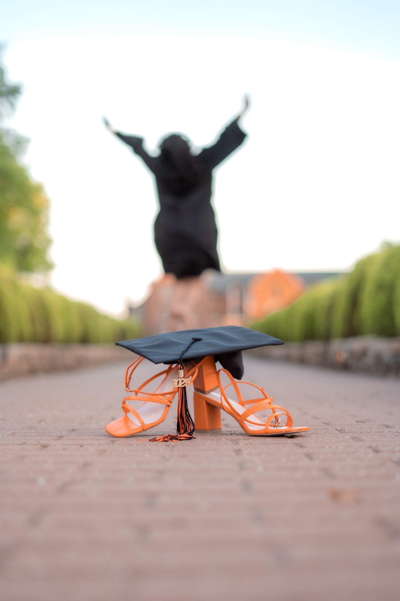 A graduation cap resting on orange heels on a brick path, with a graduate raising her arms in the distance.