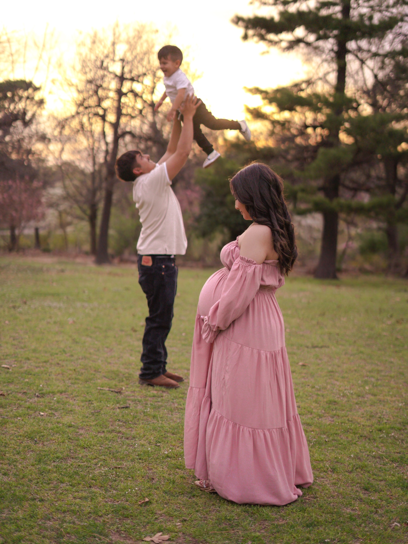 A maternity portrait at sunset: a mother in a pink dress in the foreground while a father lifts a child behind her.