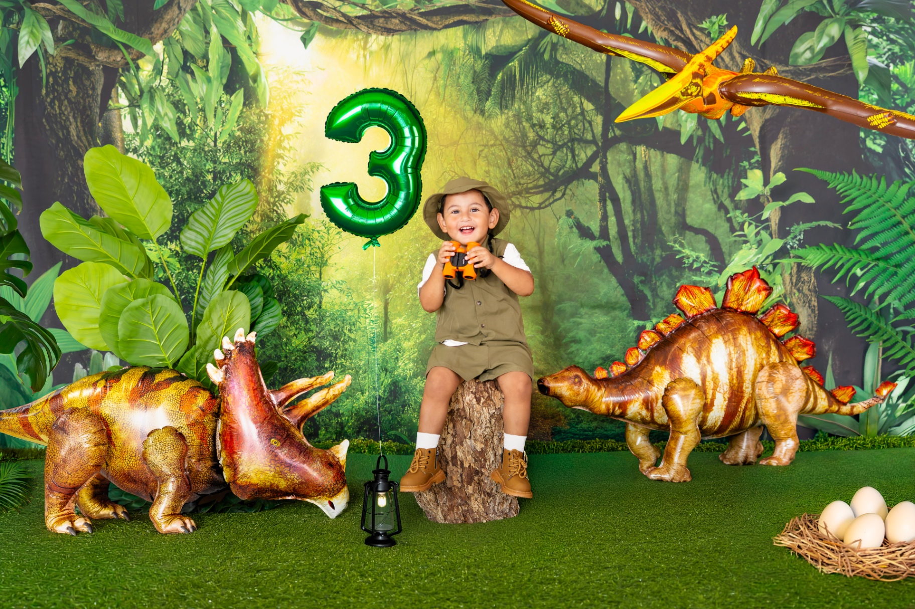 A young boy in an explorer outfit seated on a log with binoculars, surrounded by dinosaur balloons and jungle backdrop.
