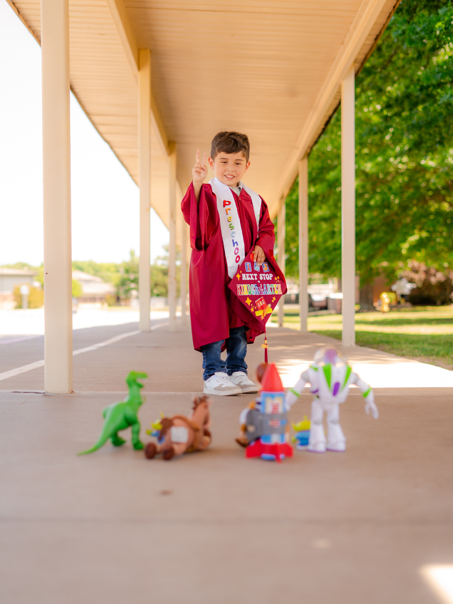 A boy in a maroon preschool graduation gown holding his cap, with toy figures on the walkway in front of him.